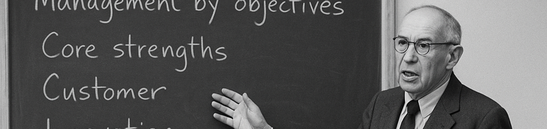 A man in a suit lectures a seated audience, chalkboard lists management topics.
